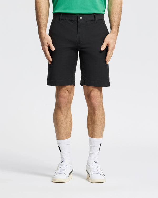 Against a plain backdrop, someone is wearing Psycho Bunny MENS FRANKLIN GOLF SHORT - B6R466F200 in black, a green shirt, white socks, and sneakers. The focus is on their lower half, highlighting the shorts' casual style.