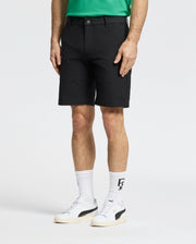 A person in a green shirt and Psycho Bunny MENS FRANKLIN GOLF SHORT - B6R466F200, with white socks and sneakers featuring black branding, stands against a plain white backdrop, highlighting the casual elegance of golf clothing mixed with athletic style. Only their lower half is visible.