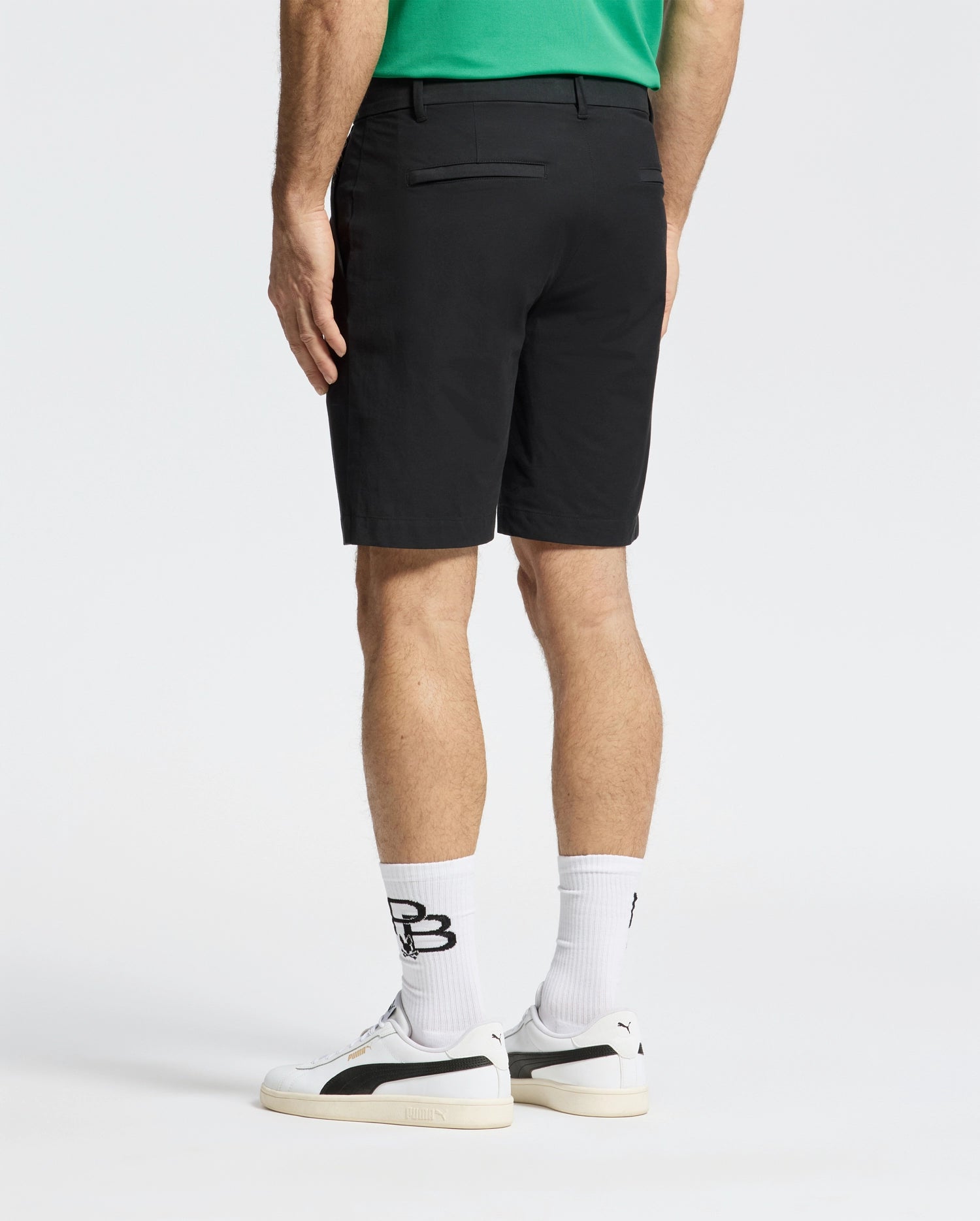 A person faces away from the camera in a green shirt, wearing Psycho Bunny's MENS FRANKLIN GOLF SHORT - B6R466F200. Their ensemble includes white socks and sneakers with black details, all against a simple, light-colored backdrop.