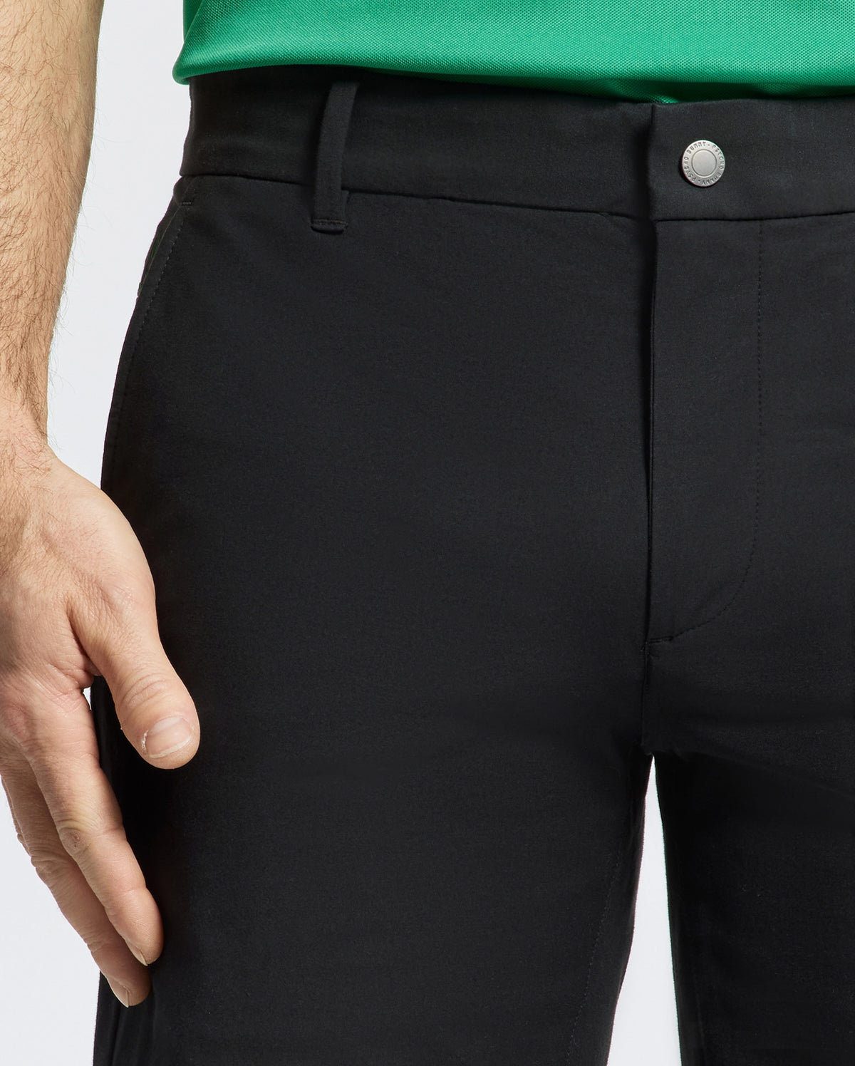 A person wearing black Psycho Bunny MENS FRANKLIN GOLF SHORT - B6R466F200 and a green shirt is shown waist down. Their left hand rests by their side, while the shorts feature belt loops and a button closure against a plain white background.