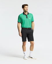 A man in a green polo with a black collar, Psycho Bunny MENS FRANKLIN GOLF SHORT - B6R466F200, white socks, and white sneakers stands against a plain backdrop.