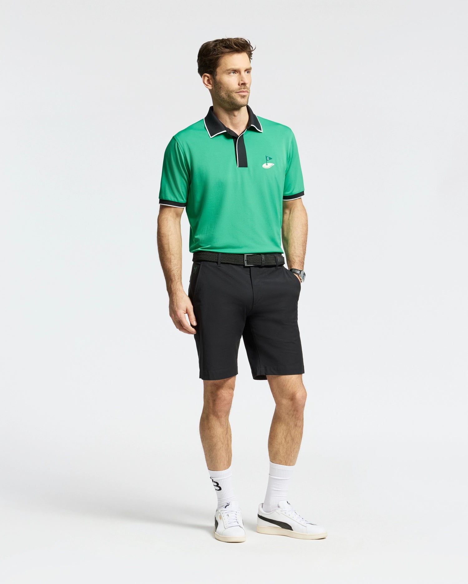 A man in a green polo with a black collar, Psycho Bunny MENS FRANKLIN GOLF SHORT - B6R466F200, white socks, and white sneakers stands against a plain backdrop.