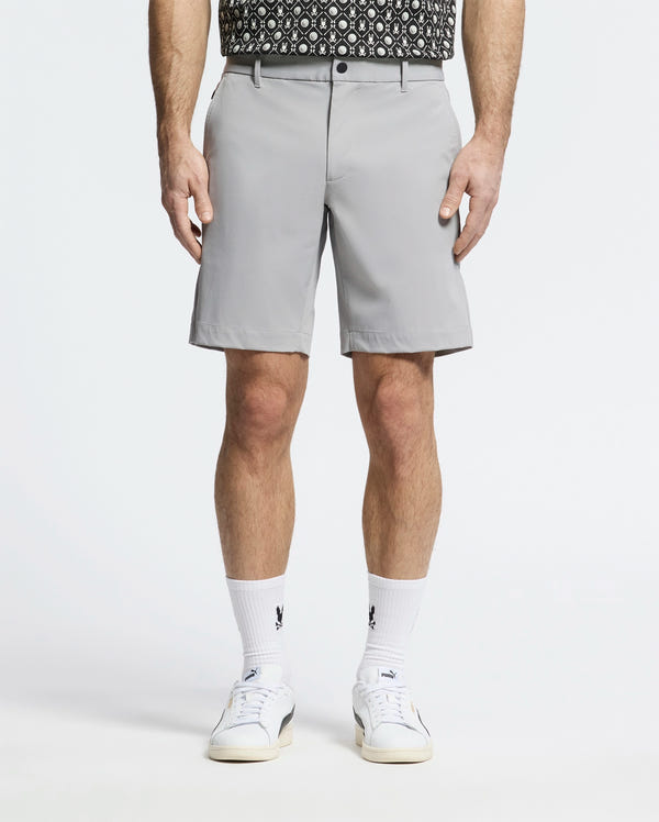 A person wearing Psycho Bunny's MENS FRANKLIN GOLF SHORT - B6R466F200 in light gray, paired with a patterned black and white shirt, logoed white socks, and white sneakers stands against a plain light background.