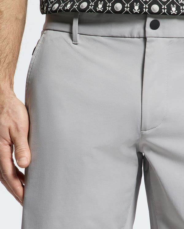 A close-up shows a person wearing Psycho Bunny's MENS FRANKLIN GOLF SHORT - B6R466F200 in light gray, paired with a black belt and a patterned black shirt with white designs partially visible at the top.
