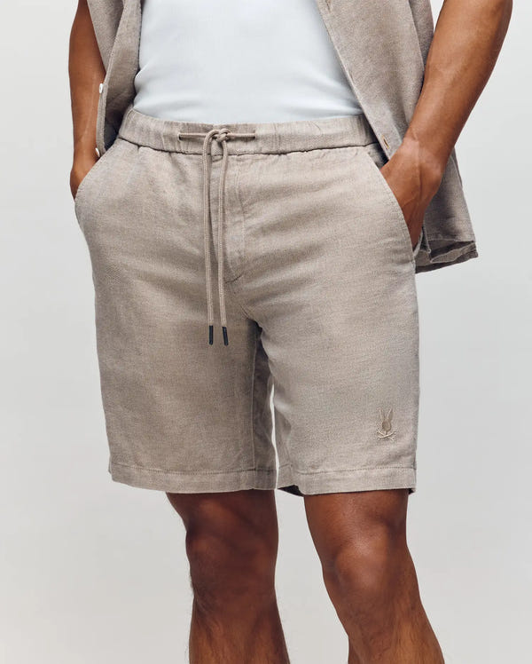 A man wears Psycho Bunny MENS WINDCREST LINEN BLEND DRAWSTRING SHORT (B6R474C200) with hands in the pockets, paired with a matching short-sleeve shirt and white top; only his lower torso and upper legs are visible.