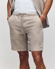 A man wears Psycho Bunny MENS WINDCREST LINEN BLEND DRAWSTRING SHORT (B6R474C200) with hands in the pockets, paired with a matching short-sleeve shirt and white top; only his lower torso and upper legs are visible.
