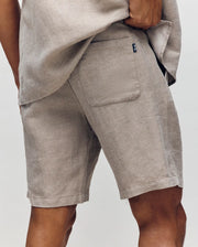 A man wears Psycho Bunny MENS WINDCREST LINEN BLEND DRAWSTRING SHORT - B6R474C200, shown waist down with a matching shirt. The shorts have a drawstring waist, back pocket with black tag, and are set against a plain background.