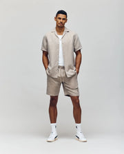 A man stands against a plain background wearing a light beige short-sleeve button-up, a white t-shirt, white socks, white sneakers, and Psycho Bunny MENS WINDCREST LINEN BLEND DRAWSTRING SHORT - B6R474C200 with a drawstring waist, hands in his pockets.