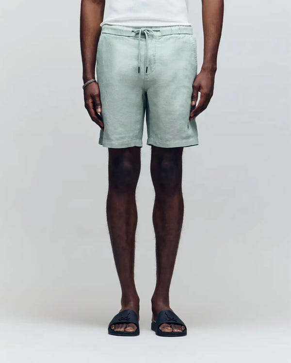 A person models the Psycho Bunny MENS WINDCREST LINEN BLEND DRAWSTRING SHORT - B6R474C200 in light green, paired with a white sleeveless shirt and black slide sandals, standing against a plain light background.