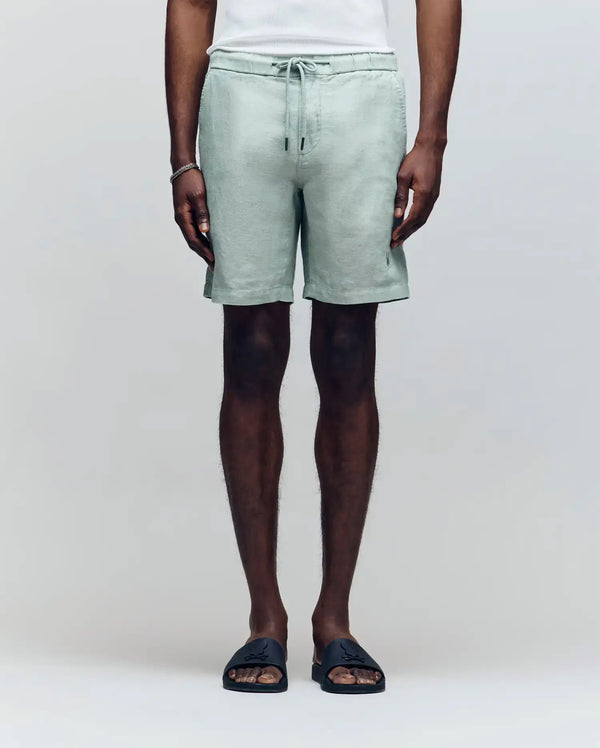 A person models the Psycho Bunny MENS WINDCREST LINEN BLEND DRAWSTRING SHORT - B6R474C200 in light green, paired with a white sleeveless shirt and black slide sandals, standing against a plain light background.