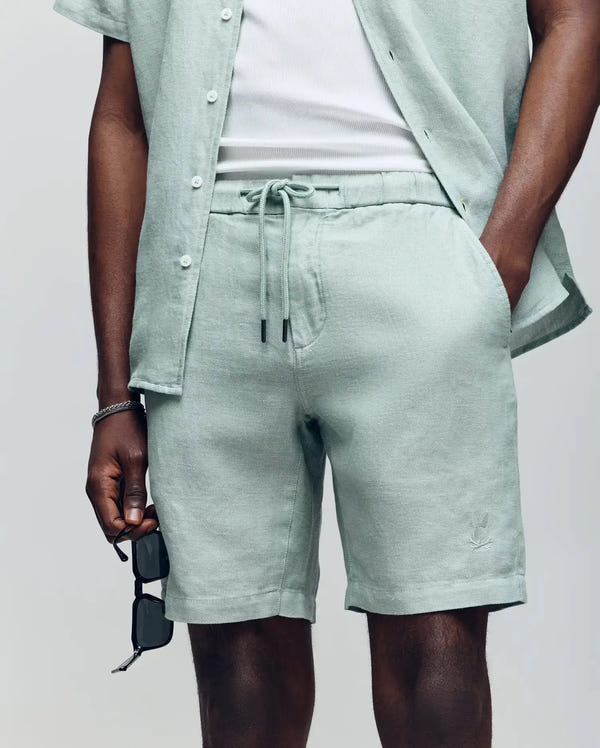 Wearing Psycho Bunny’s light green short-sleeve button-up, a white undershirt, and MENS WINDCREST LINEN BLEND DRAWSTRING SHORT - B6R474C200, a person holds black sunglasses in one hand with the other in their pocket.