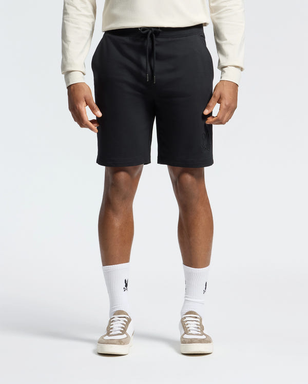 Wearing black Psycho Bunny Levanzo Embroidered Micro French Terry Sweatshorts (B6R578F200), a light long-sleeve shirt, and logo white socks exudes comfort and style. Beige sneakers complete the look against a plain, light background. The fabric ensures a soft, relaxed all-day fit.