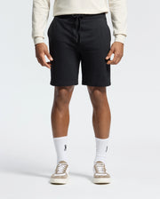 Wearing black Psycho Bunny Levanzo Embroidered Micro French Terry Sweatshorts (B6R578F200), a light long-sleeve shirt, and logo white socks exudes comfort and style. Beige sneakers complete the look against a plain, light background. The fabric ensures a soft, relaxed all-day fit.