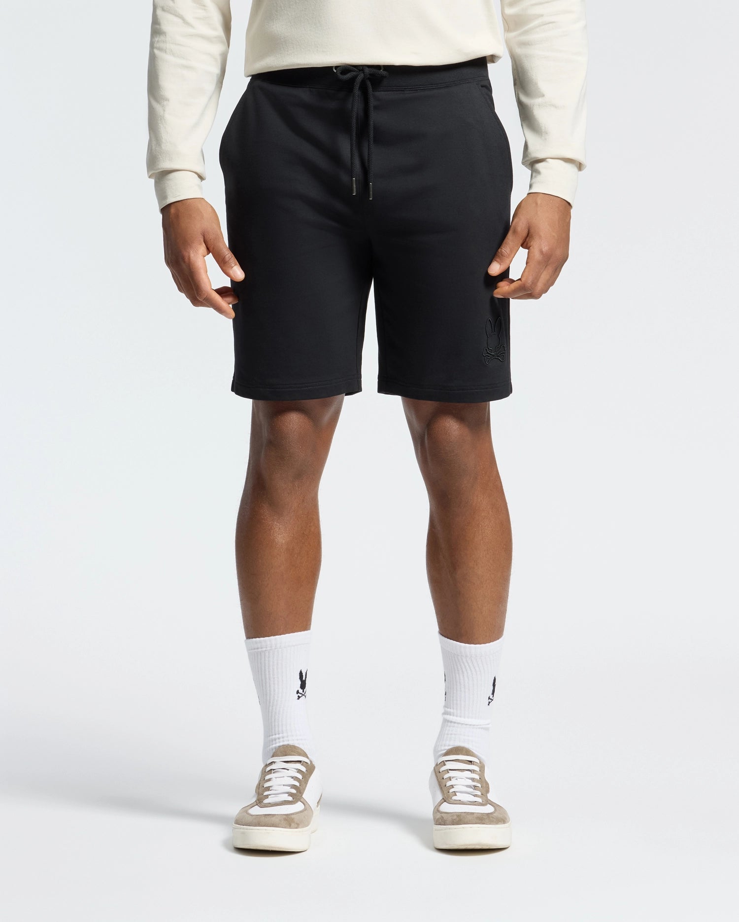 Wearing black Psycho Bunny Levanzo Embroidered Micro French Terry Sweatshorts (B6R578F200), a light long-sleeve shirt, and logo white socks exudes comfort and style. Beige sneakers complete the look against a plain, light background. The fabric ensures a soft, relaxed all-day fit.