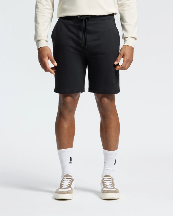 Wearing black Psycho Bunny Levanzo Embroidered Micro French Terry Sweatshorts (B6R578F200), a light long-sleeve shirt, and logo white socks exudes comfort and style. Beige sneakers complete the look against a plain, light background. The fabric ensures a soft, relaxed all-day fit.