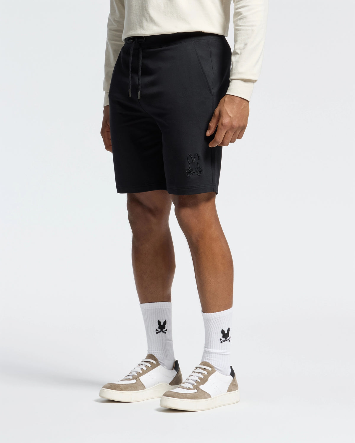 A man in a cream long-sleeve shirt, Psycho Bunny's Levanzo Embroidered Micro French Terry Sweatshorts, and white socks with an abstract rabbit logo stands against a white background. His outfit, paired with white and beige sneakers, exudes effortless comfort.