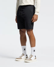 A man in a cream long-sleeve shirt, Psycho Bunny's Levanzo Embroidered Micro French Terry Sweatshorts, and white socks with an abstract rabbit logo stands against a white background. His outfit, paired with white and beige sneakers, exudes effortless comfort.