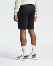 A person sports a light long-sleeve shirt and Psycho Bunny's Levanzo Embroidered Micro French Terry Sweatshort in black. Complemented by white socks and gray sneakers, this look is perfect for casual settings. The plain white background enhances the outfit’s relaxed vibe.