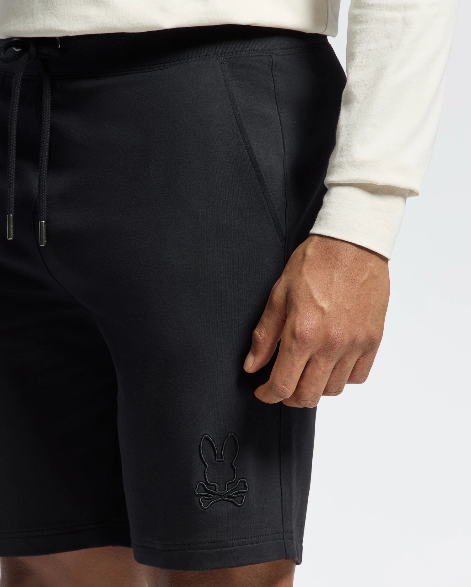 A person wearing a white long-sleeve shirt with Psycho Bunny's Levanzo Black Sweatshorts, showcasing a rabbit and crossbones logo on the leg. These Micro French Terry shorts come with drawstrings and side pockets, ensuring comfort and style.