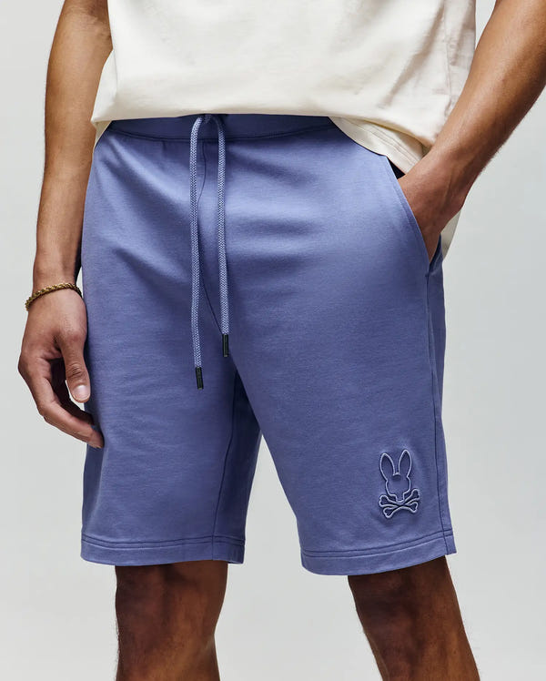 A person wears Psycho Bunny’s MENS LEVANZO FRENCH TERRY SWEATSHORT (B6R578F200) in light purple with a raised bunny and crossbones logo on the left leg, paired with a white T-shirt. Only torso and legs are visible; hands in shorts’ pockets.