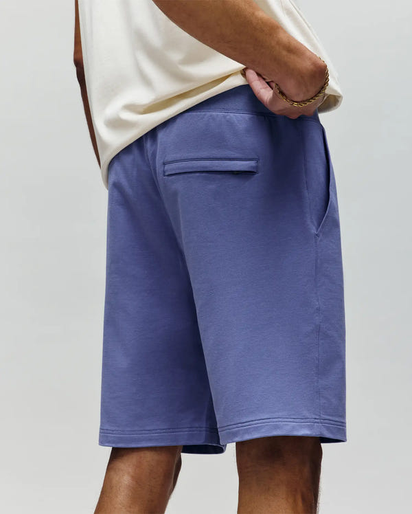 A person stands facing away, wearing a cream short-sleeve shirt and Psycho Bunny MENS LEVANZO FRENCH TERRY SWEATSHORT - B6R578F200 in light purple; the left hand rests in the pocket, with a bracelet on the right wrist.