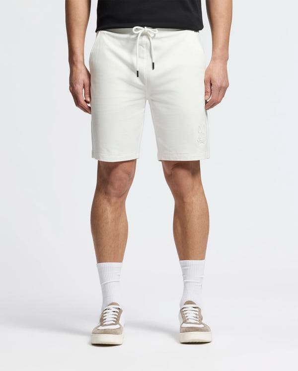 A person is wearing Psycho Bunny's Levanzo French Terry Sweatshorts in white with a bunny logo on the left leg, paired with white crew socks, beige and white sneakers, and a black top. The photo focuses on their lower half against a plain background.