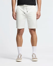 A person is wearing Psycho Bunny's Levanzo French Terry Sweatshorts in white with a bunny logo on the left leg, paired with white crew socks, beige and white sneakers, and a black top. The photo focuses on their lower half against a plain background.
