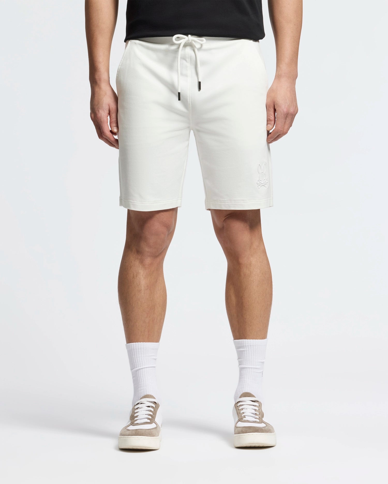 A person is wearing Psycho Bunny's Levanzo French Terry Sweatshorts in white with a bunny logo on the left leg, paired with white crew socks, beige and white sneakers, and a black top. The photo focuses on their lower half against a plain background.
