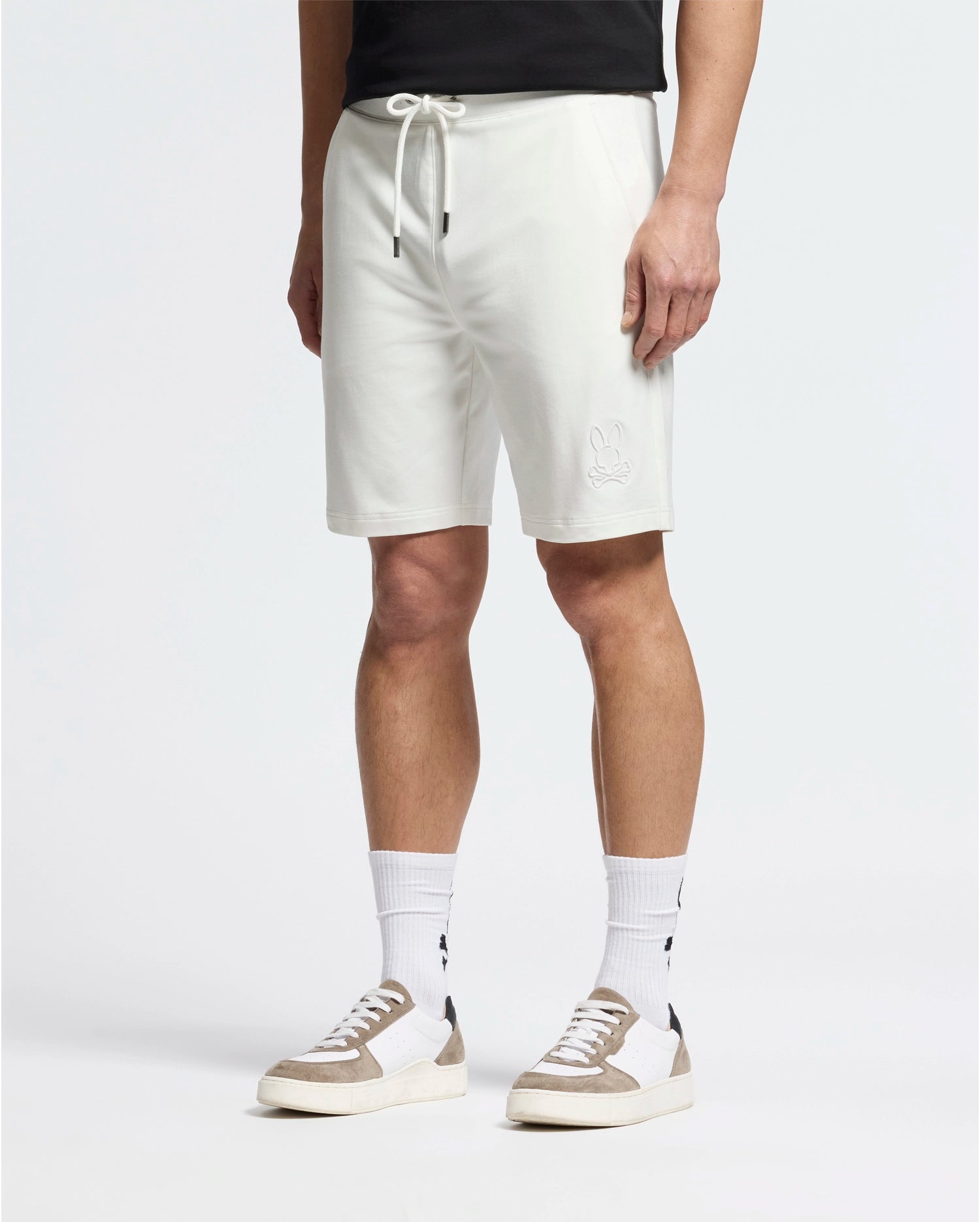 Dressed in Psycho Bunny's white Levanzo Embroidered Micro French Terry Sweatshorts featuring a bunny detail, the person pairs them with a black t-shirt, white socks with a black pattern, and sleek white sneakers accented by tan, all against a plain white background.