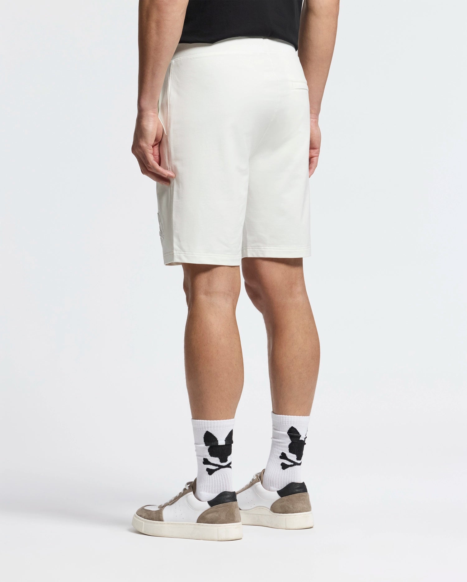 A person stands on a light background wearing a black t-shirt, white Psycho Bunny Levanzo Embroidered Micro French Terry Sweatshorts, and white socks with black skull designs. The look is finished with white sneakers featuring gray accents.