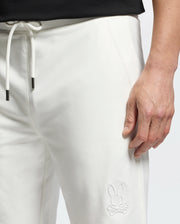 A person dons Psycho Bunny's white MENS LEVANZO EMBROIDERED MICRO FRENCH TERRY SWEATSHORT featuring a bunny with crossbones logo. A black drawstring peeks at the waistband as they hold one hand by their side. The texture contrasts with their black top.