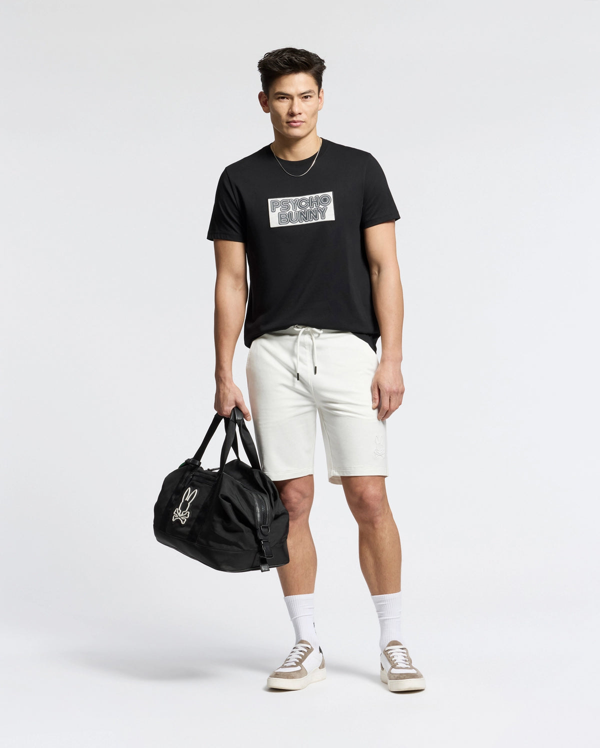 A person in a Psycho Bunny black tee with an embroidered bunny graphic and MENS LEVANZO EMBROIDERED MICRO FRENCH TERRY SWEATSHORT - B6R578F200 holds a black duffel bag against a plain white background, pairing it with white socks and cream sneakers.