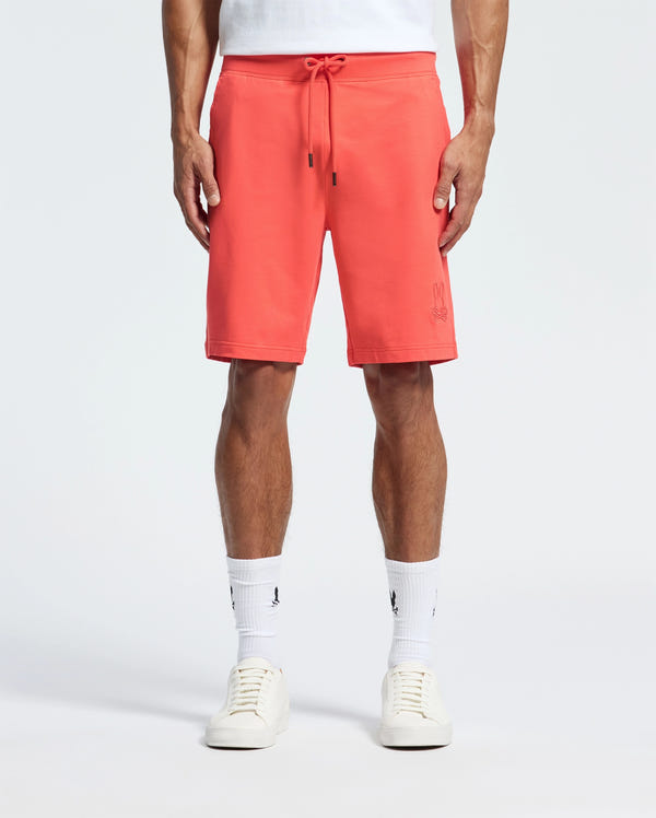 A person wears coral pink Psycho Bunny Men's Levanzo French Terry Sweatshorts (B6R578F200) with a drawstring, white sneakers, and visible white crew socks against a plain background. Crafted from micro French terry for comfort, the shorts have a subtle logo on one leg.
