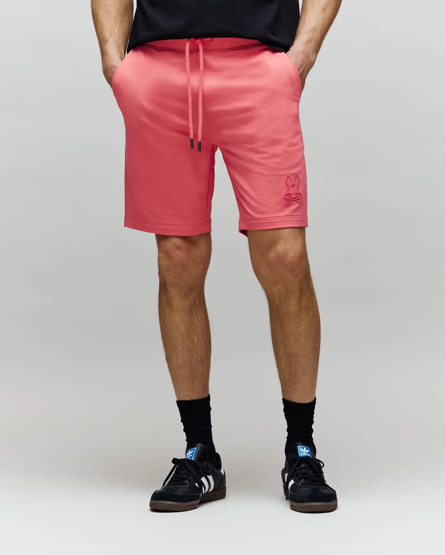 A person wears Psycho Bunny MENS LEVANZO FRENCH TERRY SWEATSHORT - B6R578F200 in bright pink, styled with a black t-shirt, black socks, and black sneakers, standing hands-in-pockets against a plain light background.