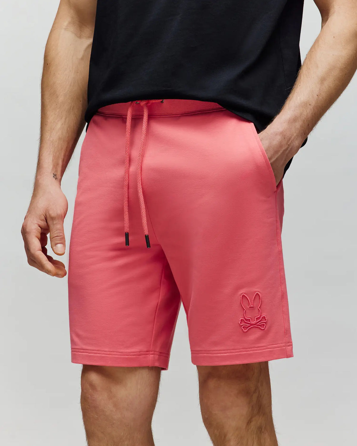 A person wears Psycho Bunny MENS LEVANZO FRENCH TERRY SWEATSHORT (B6R578F200), bright pink with a bunny and crossbones logo on the left leg, styled with a black tee. The hand-in-pocket pose shows comfort against a light gray background.