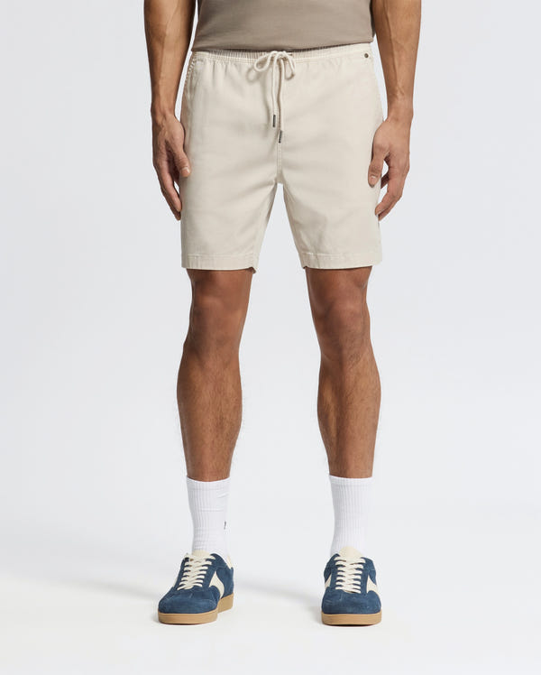 Effortlessly stylish, a person wears beige Psycho Bunny MENS WILLIS STRETCH TENCEL SHORTS (B6R584F200) with an elastic waistband, a taupe tank top, white socks, and blue sneakers against a plain white background.