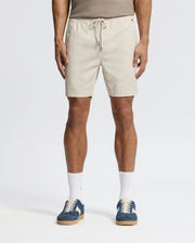 Effortlessly stylish, a person wears beige Psycho Bunny MENS WILLIS STRETCH TENCEL SHORTS (B6R584F200) with an elastic waistband, a taupe tank top, white socks, and blue sneakers against a plain white background.