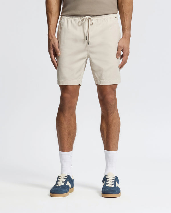 Effortlessly stylish, a person wears beige Psycho Bunny MENS WILLIS STRETCH TENCEL SHORTS (B6R584F200) with an elastic waistband, a taupe tank top, white socks, and blue sneakers against a plain white background.