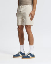 A person exudes effortless style in Psycho Bunny's light tan MENS WILLIS STRETCH TENCEL SHORT featuring an elasticized waistband and drawstring. Paired with blue sneakers featuring white accents and logo-printed white socks, they pose against a plain white background.