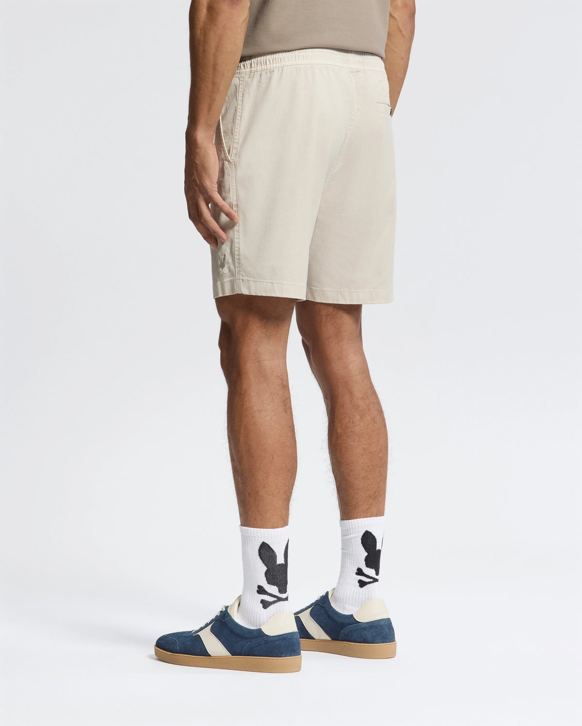 A person exudes effortless style in Psycho Bunny's MENS WILLIS STRETCH TENCEL SHORTS in beige, paired with a taupe shirt. They sport white socks with black skulls and crossbones, alongside blue and beige sneakers. A plain white backdrop highlights their laid-back vibe.