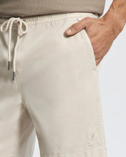 A close-up showcases a person wearing Psycho Bunny's MENS WILLIS STRETCH TENCEL SHORT - B6R584F200 in beige, featuring an elasticized waistband and hands-in-pockets style. An embroidered logo adorns the hem against a plain white backdrop.