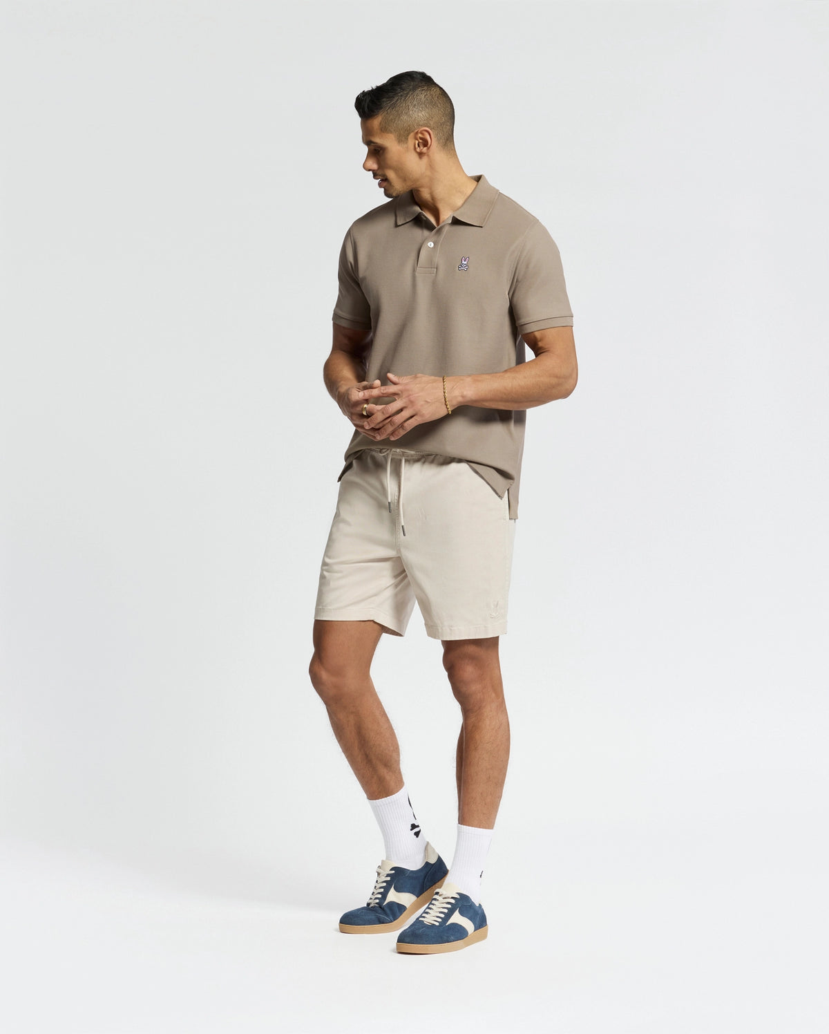 Wearing a tan polo shirt, Psycho Bunny's Men's Willis Stretch Tencel Shorts in light beige with an elasticized waistband, white socks, and blue sneakers, a man stands stylishly against a plain white background, gazing right with his hands at his sides.