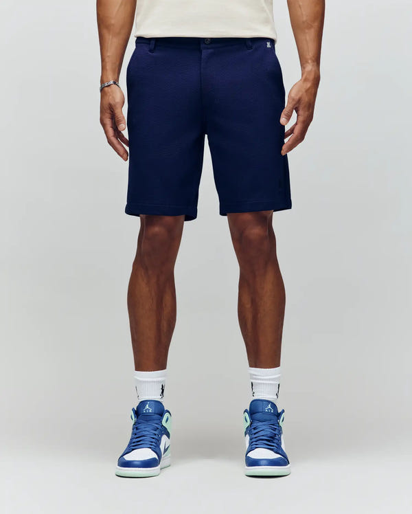 A person showcases Psycho Bunny's MENS SHIRO KNIT HONEYCOMB SHORT - B6R585F200 in navy blue, paired with white socks and blue-white sneakers, standing against a light gray background with the embroidered Bunny logo in focus.