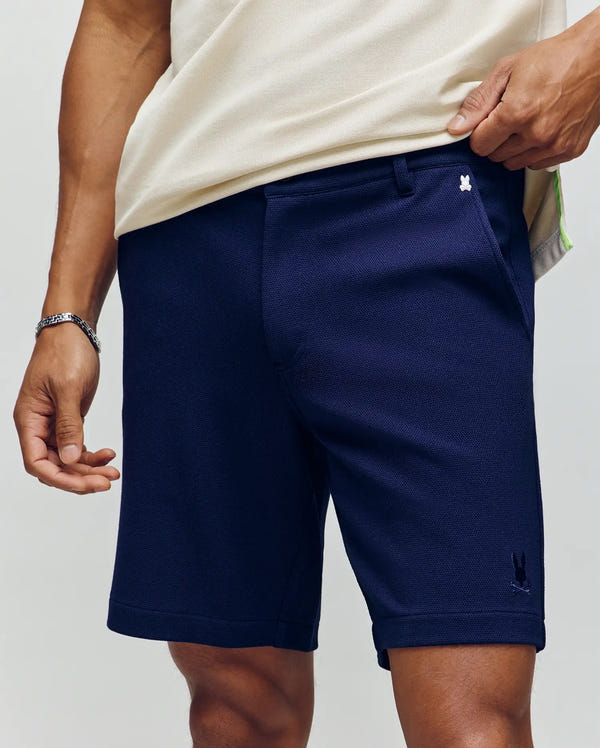 A person wears a cream-colored shirt and Psycho Bunny MENS SHIRO KNIT HONEYCOMB SHORT - B6R585F200 in dark navy, featuring an embroidered Bunny near the pocket, standing with one arm relaxed by their side.