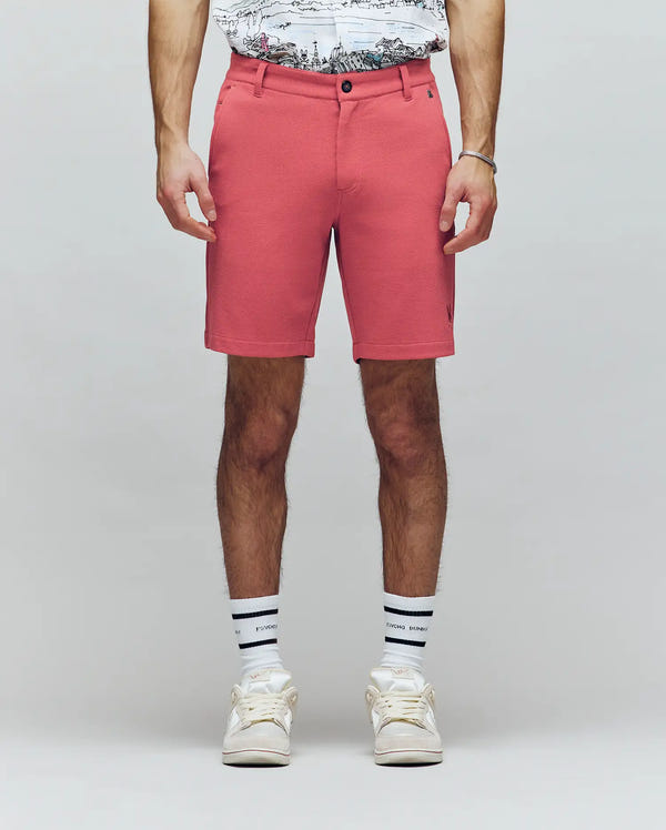 A person wearing Psycho Bunny MENS SHIRO KNIT HONEYCOMB SHORT - B6R585F200 in coral, a honeycomb piqué T-shirt, white socks with black stripes, and white sneakers stands against a plain light background. Only the lower half is visible.