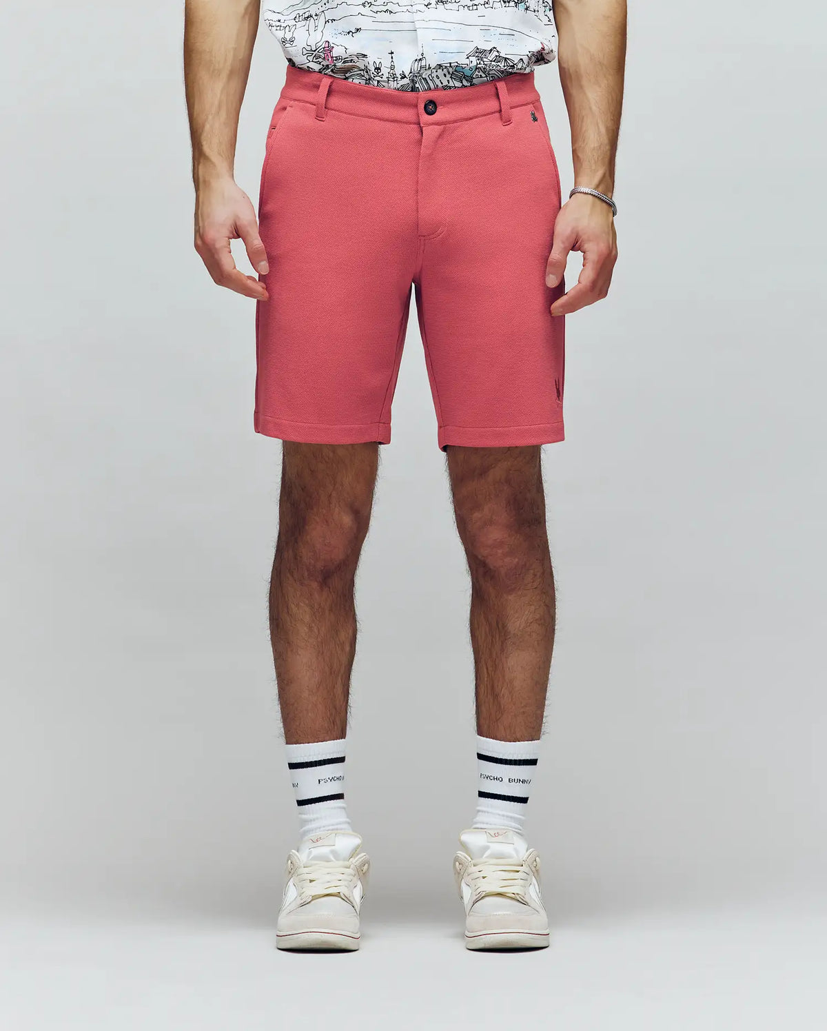A person wearing Psycho Bunny MENS SHIRO KNIT HONEYCOMB SHORT - B6R585F200 in coral, a honeycomb piqué T-shirt, white socks with black stripes, and white sneakers stands against a plain light background. Only the lower half is visible.