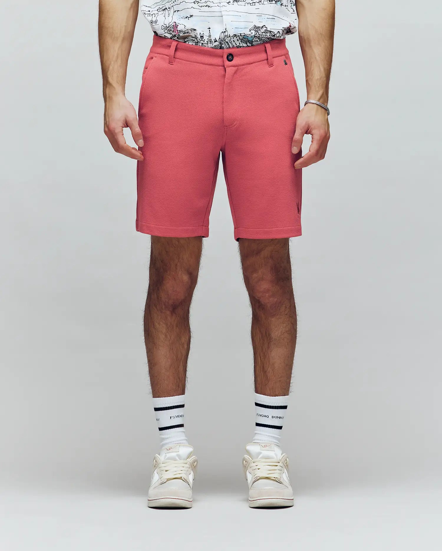 A person wearing Psycho Bunny MENS SHIRO KNIT HONEYCOMB SHORT - B6R585F200 in coral, a honeycomb piqué T-shirt, white socks with black stripes, and white sneakers stands against a plain light background. Only the lower half is visible.