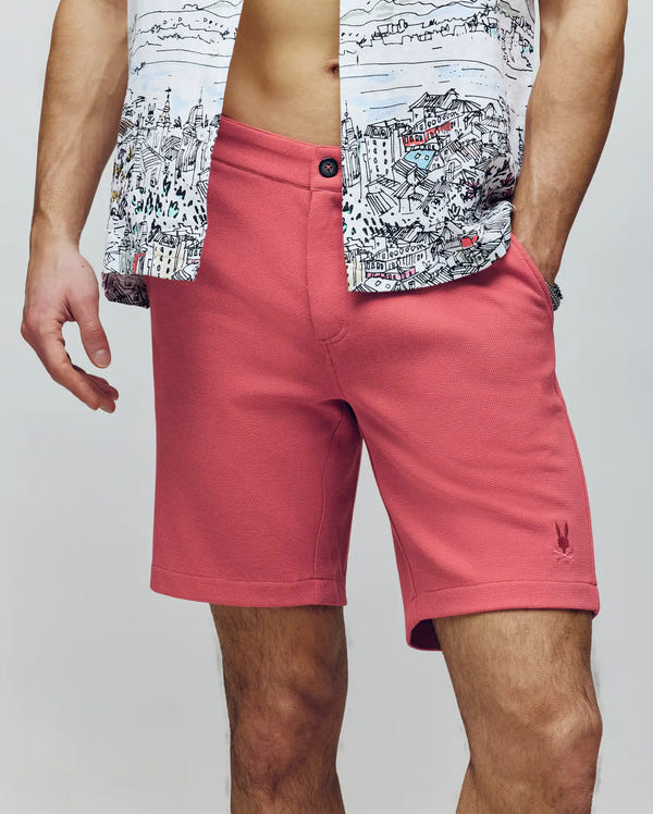 A man wears Psycho Bunny's MENS SHIRO KNIT HONEYCOMB SHORT - B6R585F200 in pink and a partially unbuttoned cityscape-print shirt, revealing his midriff. His hands rest by his sides against a plain, neutral background.
