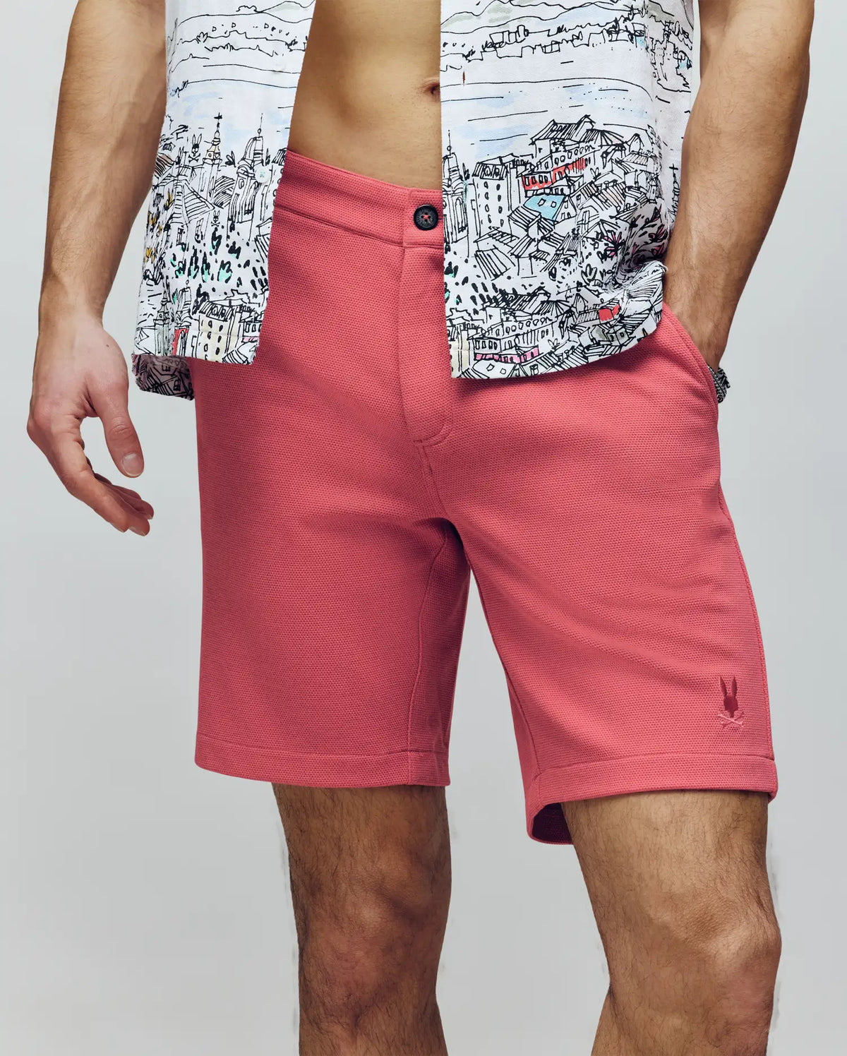 A man wears Psycho Bunny's MENS SHIRO KNIT HONEYCOMB SHORT - B6R585F200 in pink and a partially unbuttoned cityscape-print shirt, revealing his midriff. His hands rest by his sides against a plain, neutral background.