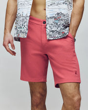 A man wears Psycho Bunny's MENS SHIRO KNIT HONEYCOMB SHORT - B6R585F200 in pink and a partially unbuttoned cityscape-print shirt, revealing his midriff. His hands rest by his sides against a plain, neutral background.